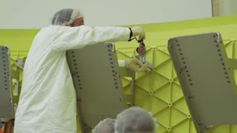 A technician in a white coat operates a tool on a green panel.