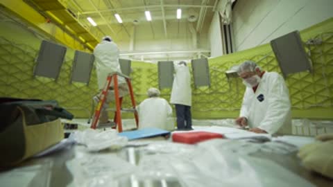 Technicians work inside a large facility with green structural panels.
