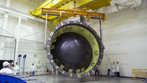 A large cylindrical structure is hoisted by overhead cranes in a facility.