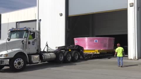 A semi-truck pulls out of a building with a pink-wrapped cylindrical component.