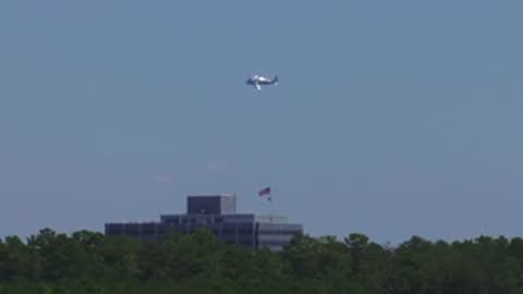 A small airplane flies across a clear blue sky over a tree line.