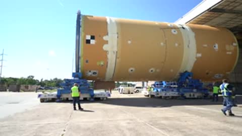 Rocket component being transported outdoors on specialized wheeled transporters by workers.
