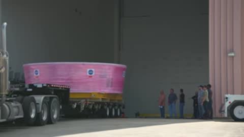 A semi-truck transports a pink-wrapped component past a building with onlookers.