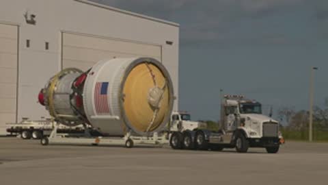 A semi-truck transports a large cylindrical rocket stage outdoors in daylight.