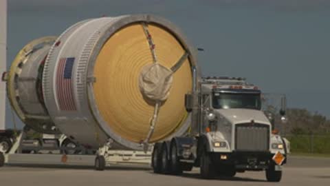 A large rocket stage is transported on a flatbed trailer by a truck.