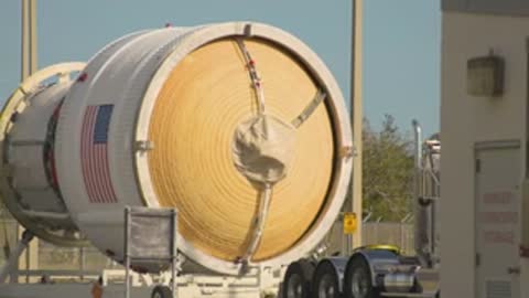 A semi-truck hauls a large cylindrical rocket stage past a fenced area.