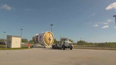 A semi-truck carrying a rocket stage navigates a turn on a paved road.