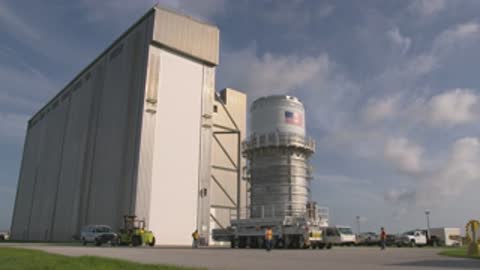 medium shot of rocket section transported past large building exterior