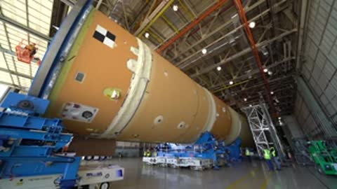 Large cylindrical rocket component on transporters inside a factory.