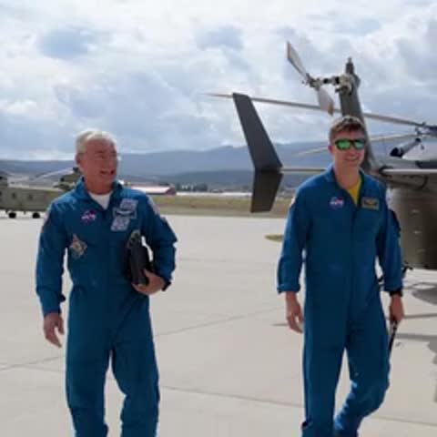 Two astronauts in blue flight suits walking outdoors near parked helicopters.