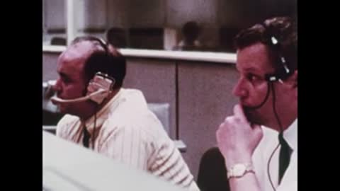 Close-up of two men wearing headsets talking closely at their station