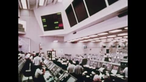 Wide angle view of a filled Mission Control room with monitors overhead