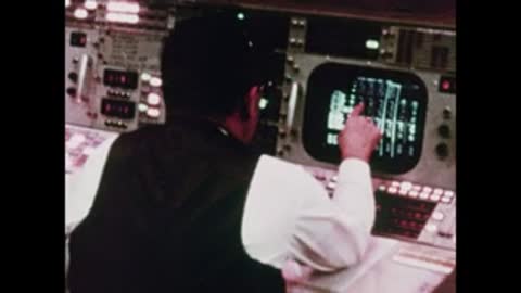 A flight controller pointing at a glowing screen on his console, back view.