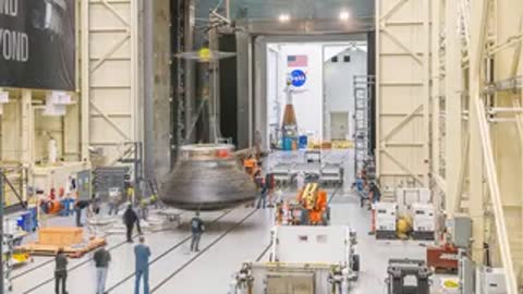 Wide shot of technicians in assembly building using crane to lift spacecraft component