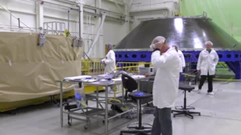 Engineers working near a rocket stage in a large assembly workshop.