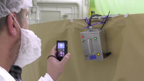 An engineer filming an electronic testing box mounted on a tent wall with a phone.