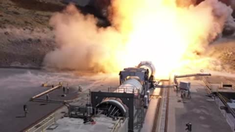 A massive rocket booster firing test igniting with huge flames and smoke in a desert landscape.