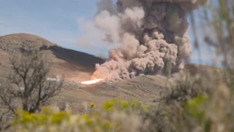 Thick brown and grey smoke rising from a rocket booster test in a hilly landscape.