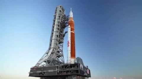 Static medium shot of SLS on crawler transporter in broad daylight.