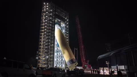 A large cylindrical tank section being lifted vertically inside a tall test tower at night.