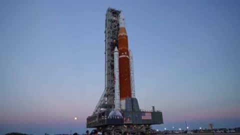 Static shot of SLS on crawler transporter at dusk under blue sky.