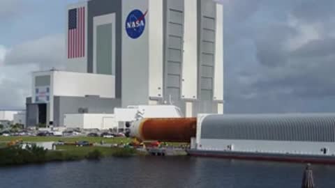The orange core stage of the SLS rocket being transported on a barge outside the VAB.