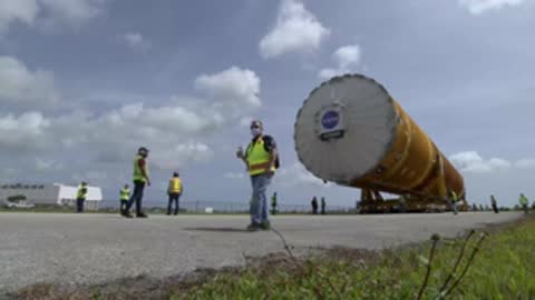 Workers in high-visibility vests walking in front of the large cylindrical core stage outdoors.