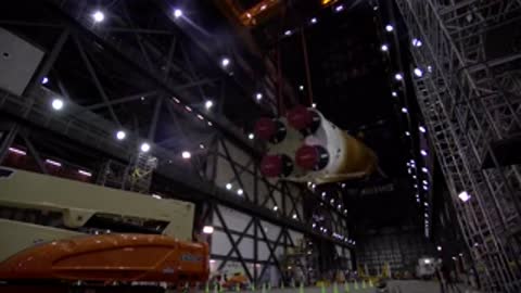 A crane lifting and tilting the massive orange core stage upright inside the assembly building.