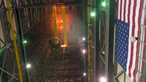 A cylindrical adapter component being lowered inside the VAB past a large American flag.
