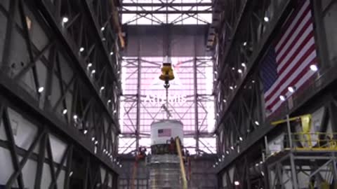 The Interim Cryogenic Propulsion Stage being lifted inside the VAB with a large American flag behind.
