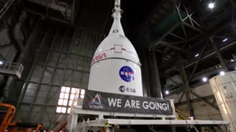 The Orion spacecraft encased in its fairing standing atop a transport platform with a banner reading WE ARE GOING.