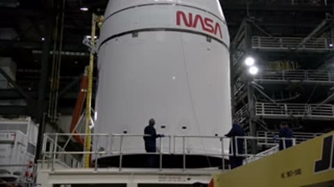 Workers standing on platforms beside the large white conical Orion spacecraft section inside the VAB.
