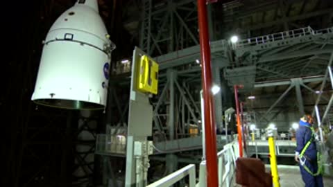 A white cylindrical rocket component is being hoisted by a crane in an assembly facility.