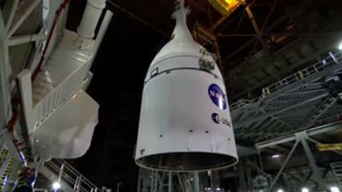 A crane lifts a large white rocket segment inside a tall industrial facility.