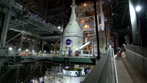 A tall rocket assembly stands vertically inside a brightly lit manufacturing facility.
