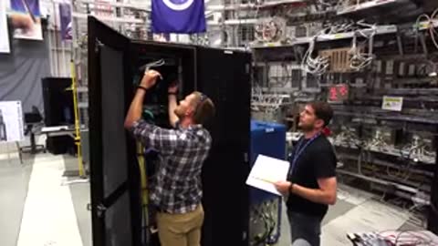 A man opens a server rack door while holding a clipboard in a facility.