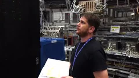 A man holding a clipboard looks up intently inside a facility with server racks.