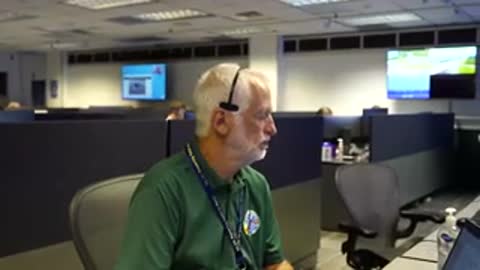 An operator with a headset speaks while sitting at a desk in a control room.