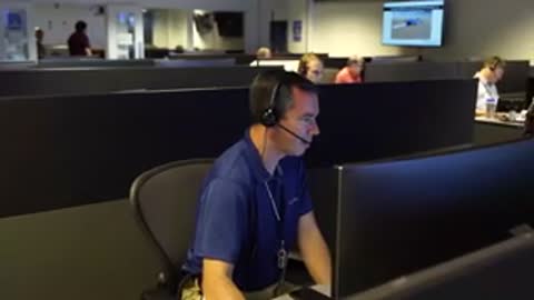 An operator in a blue shirt sits at a computer terminal in a control room.