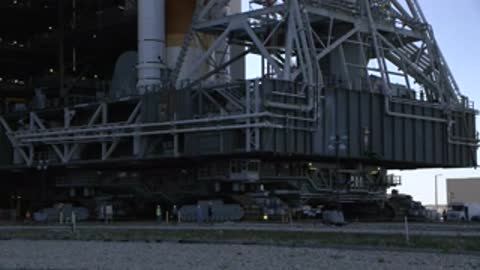 Low-angle tracking shot of crawler transporter treads moving across ground.