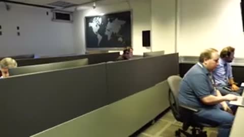 Operators sit at desks in a control room with a world map on the wall.