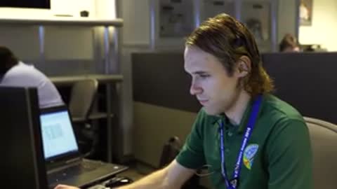 A young operator in a green polo shirt works at a computer in a control room.