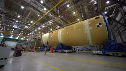 large cylindrical rocket stage resting horizontally on blue transporter inside factory building