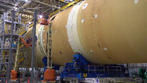 workers on lift platforms working on massive cylindrical rocket stage inside manufacturing facility