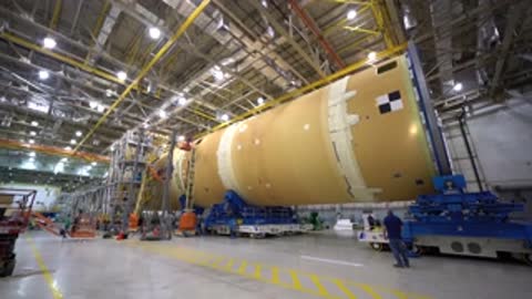 wide view of large cylindrical rocket stage in assembly building with workers and equipment