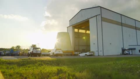 transport vehicle carrying conical rocket adapter approaching large hangar building outdoors
