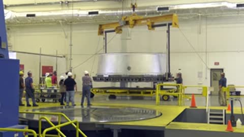 workers gathered around large cylindrical adapter suspended by crane in assembly building