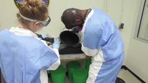 People in lab coats and safety gear handle dark material at a table.