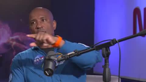 Speaker in blue flight suit gesturing during interview against blue background.
