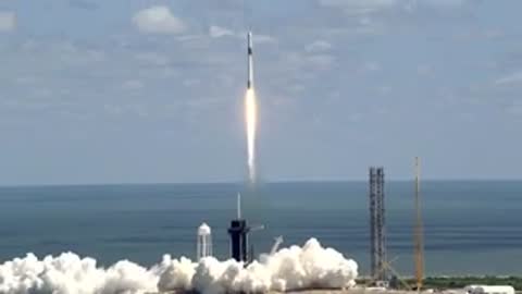 Rocket launching from pad near water into cloudy sky.